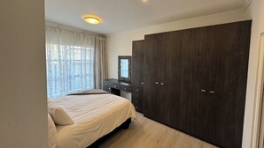 A bedroom with a bed, a dark wooden wardrobe, a dresser, and a window with curtains. - 2BR-2BA - Rivonia Residence 3 (Sandton)