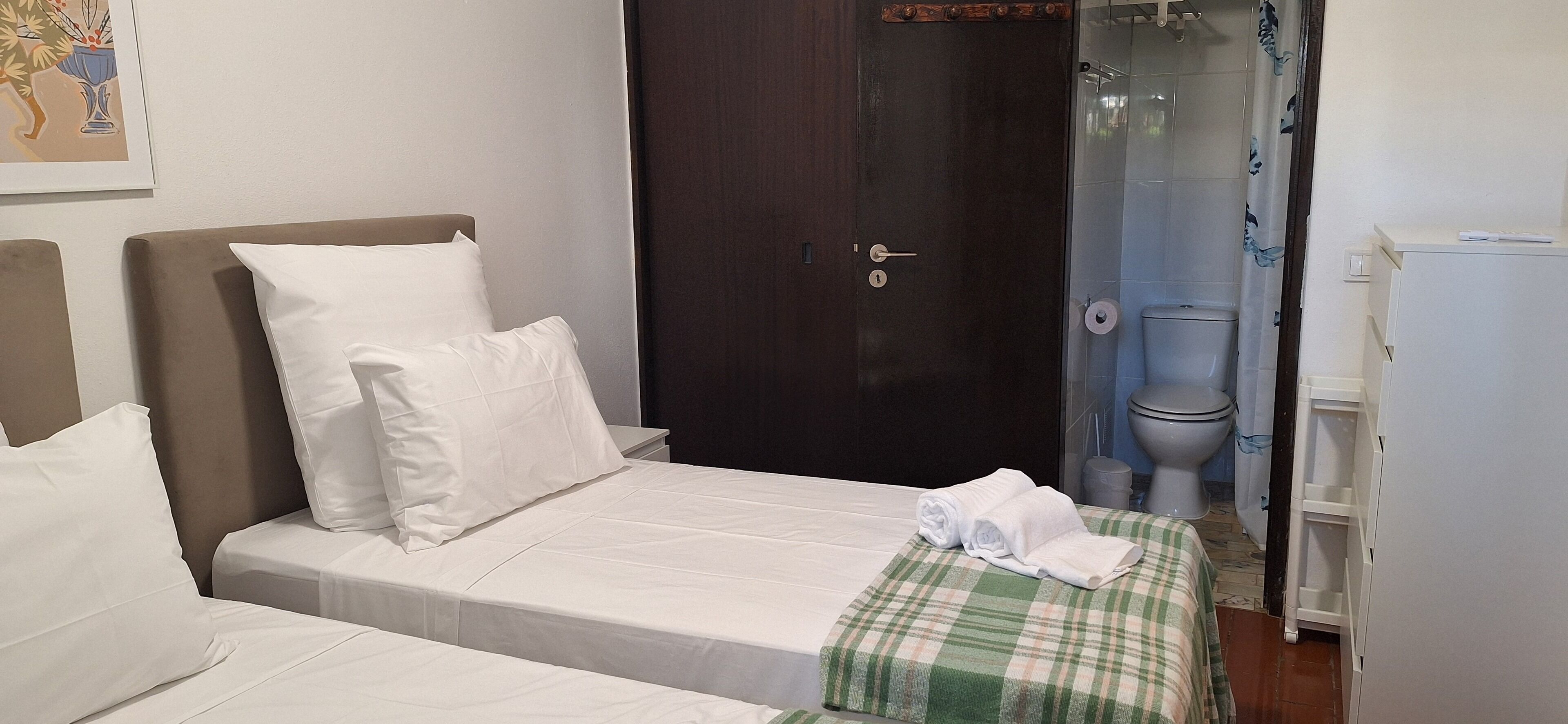 Ein Hotelzimmer mit zwei Betten, einer dunklen Holztür, einer Dusche und einem weißen Kühlschrank.