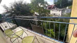 A balcony with metal railings, a chair, and a view of a tree and buildings in the background.