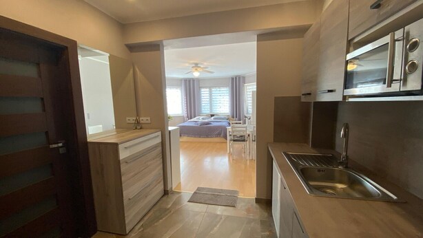 A modern kitchen with a built-in oven and sink, wooden cabinets, and a view into a bedroom with a bed and curtains.