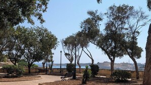 Un parque pavimentado con bancos y árboles, un farol y vistas al mar.