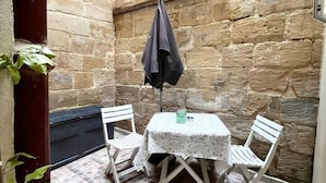 Un pequeño patio con pared de piedra, suelo de baldosas, una mesa con mantel de encaje, dos sillas blancas y una chaqueta oscura colgada.