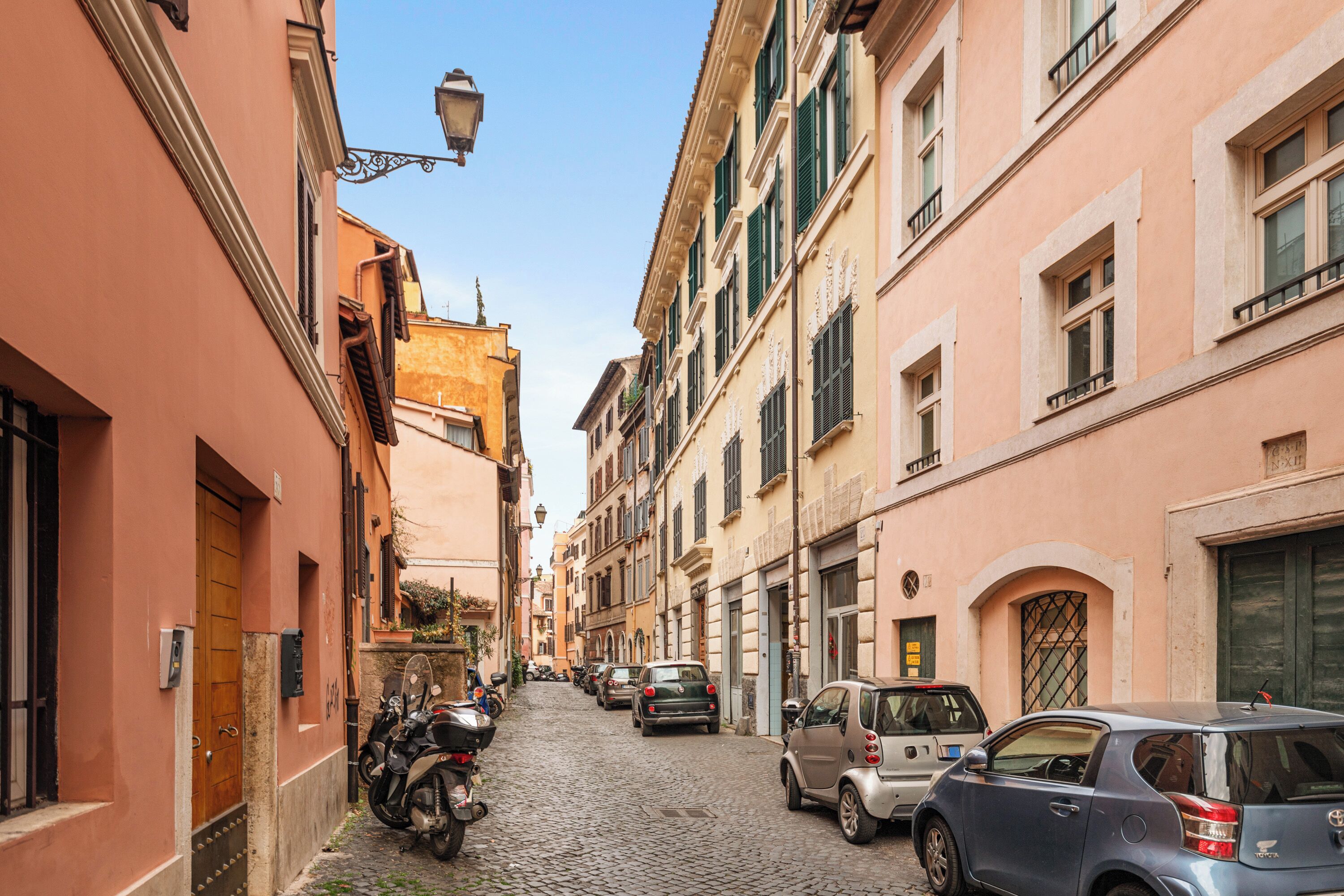 A narrow cobblestone street lined with historic buildings, parked cars, and motorcycles.