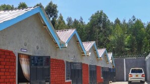 A row of small, single-story buildings with red brick walls and blue roofs, separated by a high concrete wall with barbed wire. - Paradise Point Resort Naivasha (Nyahururu)