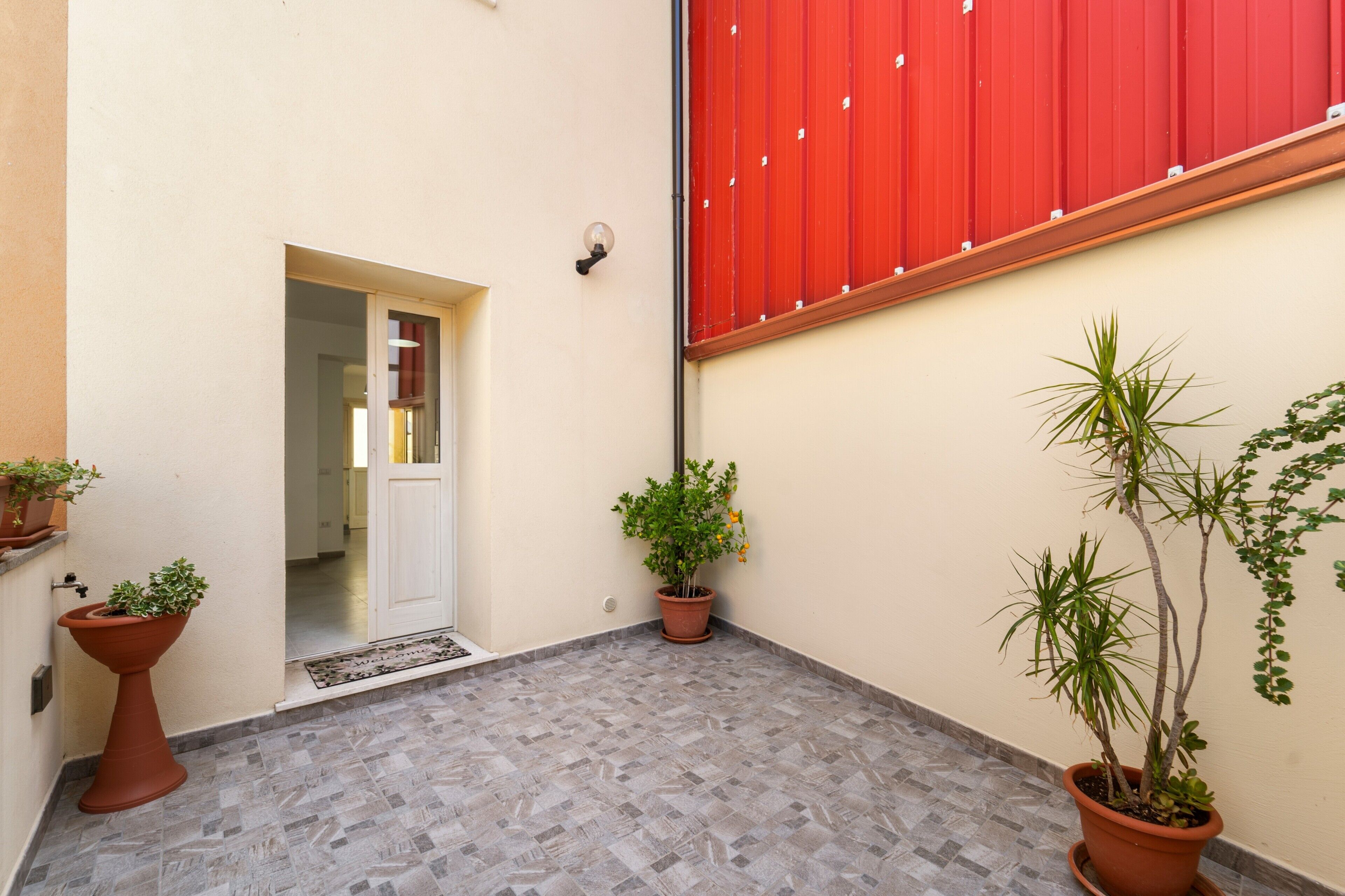 Un couloir étroit au sol carrelé, agrémenté de plantes en pot, avec une porte rouge et un luminaire fixé au mur.