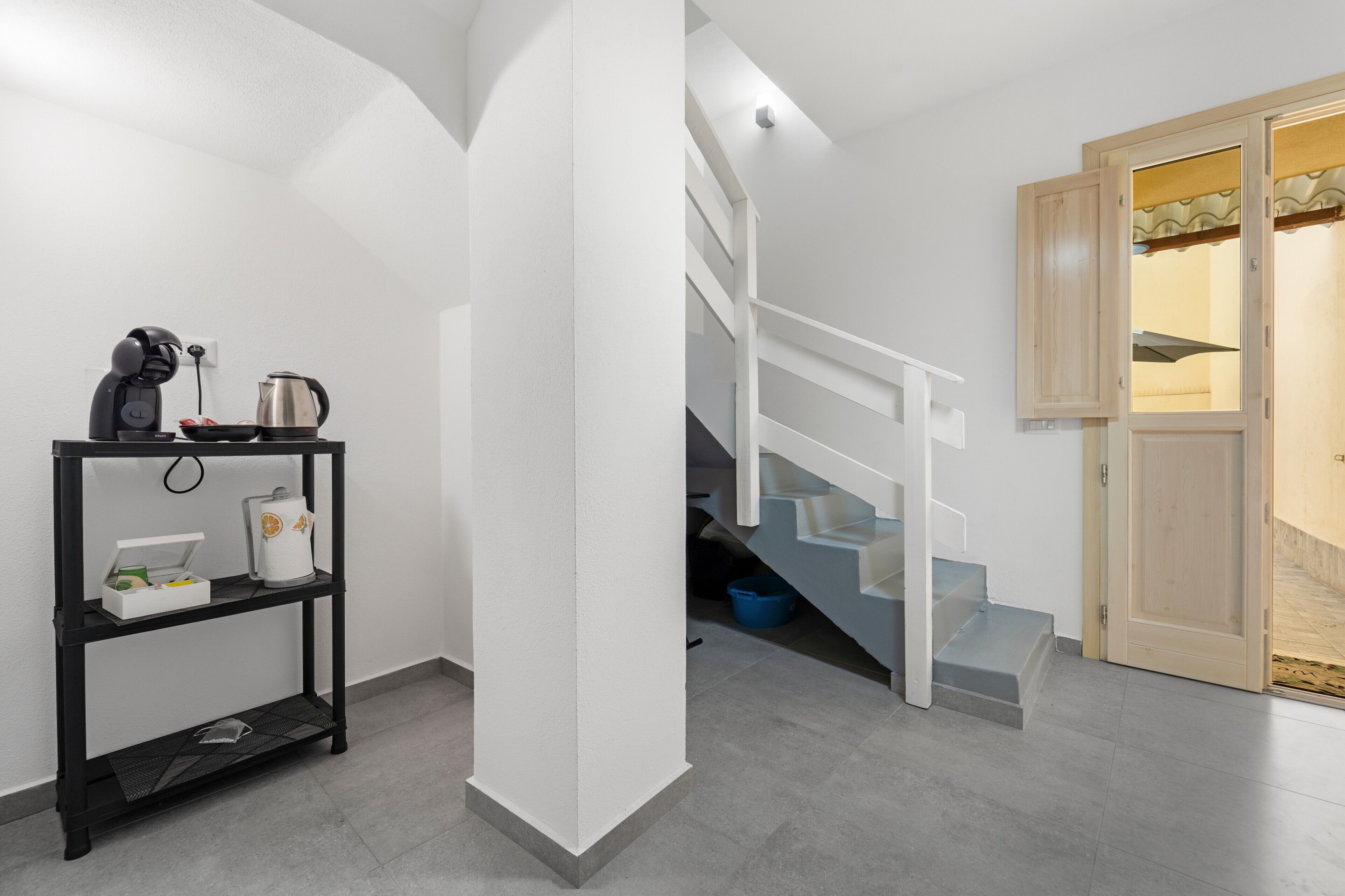 Un escalier menant à un deuxième étage, une étagère en métal noir contenant des ustensiles de cuisine et une porte en bois donnant sur un paysage agréable.