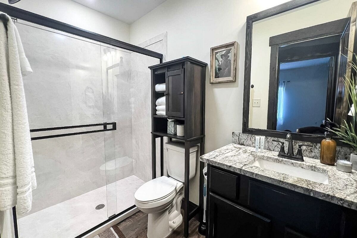 A bathroom with a glass-enclosed shower, a toilet, a dark cabinet with towels, a marble sink, and a large mirror.