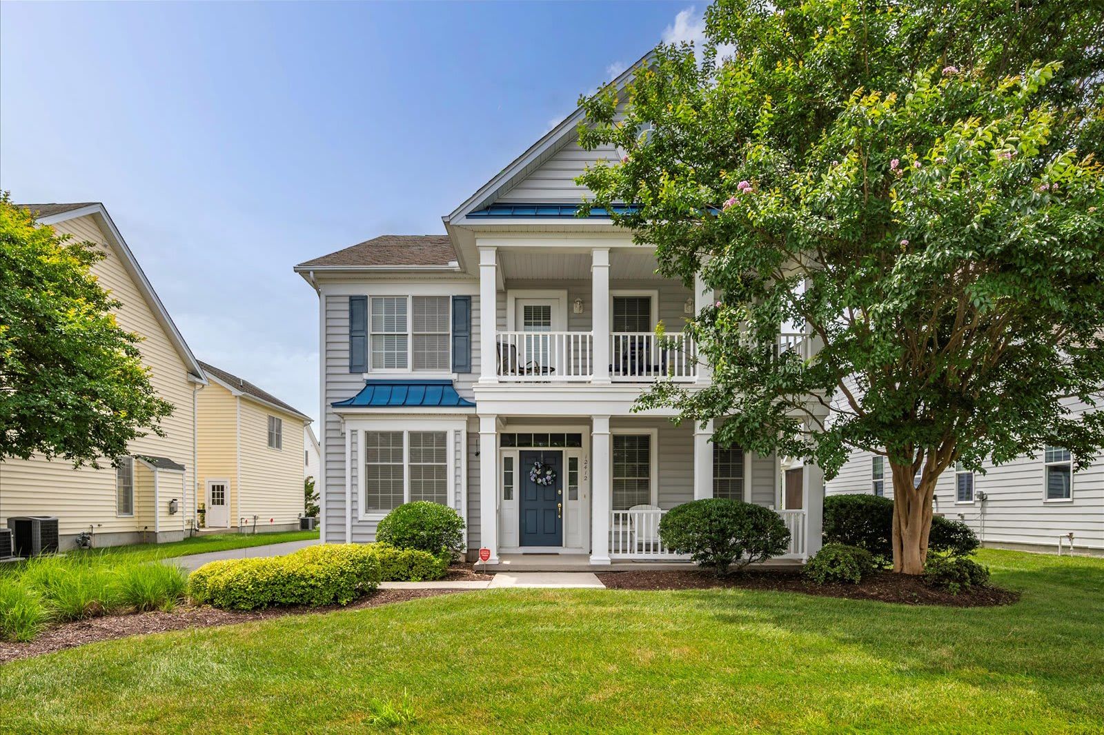 A two-story residential building with a front porch, a small front yard, and a tree.