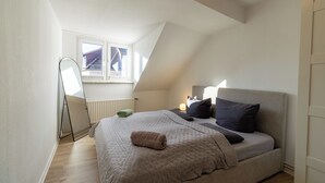 A bedroom with a sloped ceiling, a bed with a beige quilt, a bedside lamp, and a window with a view of buildings.
