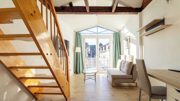 A loft-style bedroom with a wooden staircase, a sofa, a desk, and a view through a glass door.