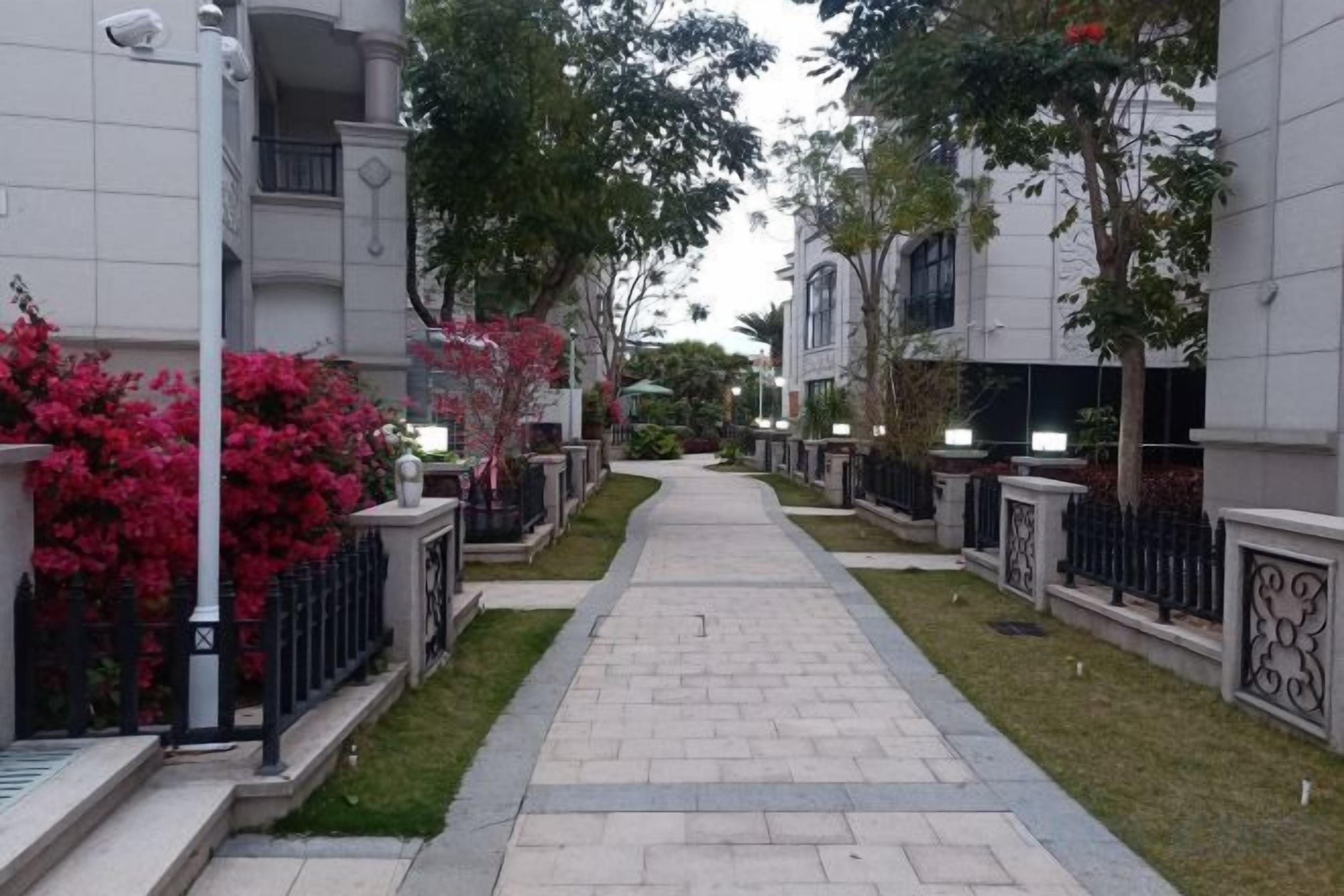 A pathway flanked by buildings with balconies, surrounded by greenery and decorative fencing.