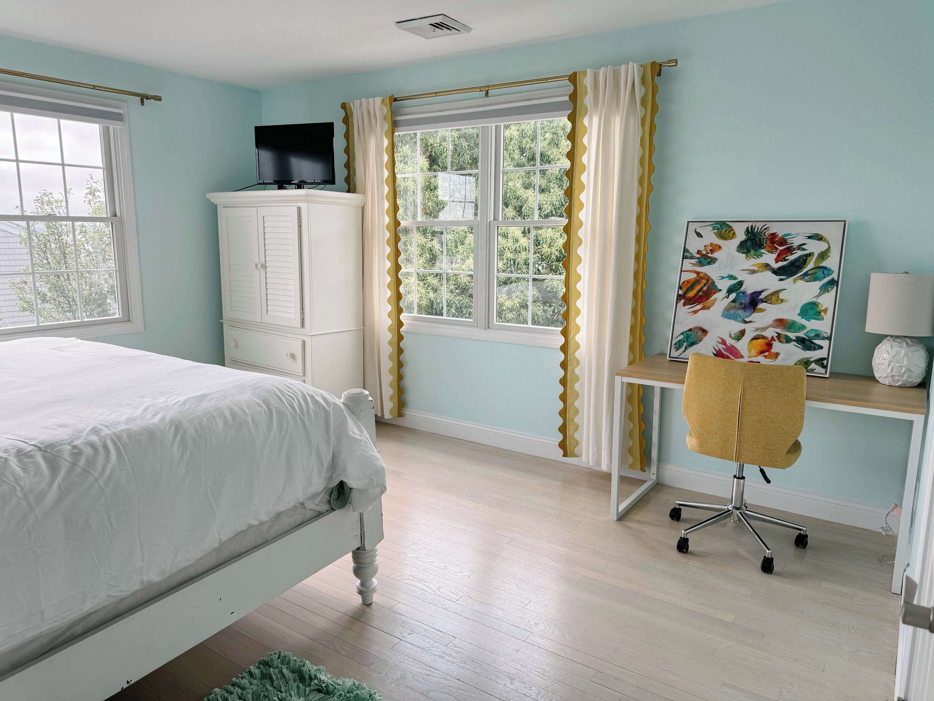A bedroom with a bed, a desk with a chair, a TV on a cabinet, and a window with curtains.