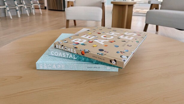 Een stapel boeken met de titel 'Beaches, Coastal, Escape' van Gray Malin op een ronde houten tafel in een woonkamer met een modern design, inclusief een bank en stoelen.