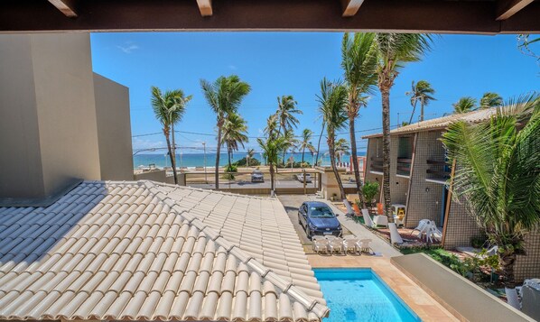 A swimming pool with a tiled roof, a view of the ocean, palm trees, and a building with balconies.