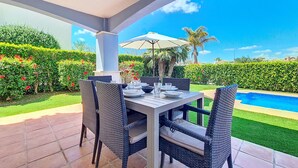 Um pátio externo com mesa posta para refeições, rodeado por cadeiras de vime, uma piscina e sebes bem cuidadas.