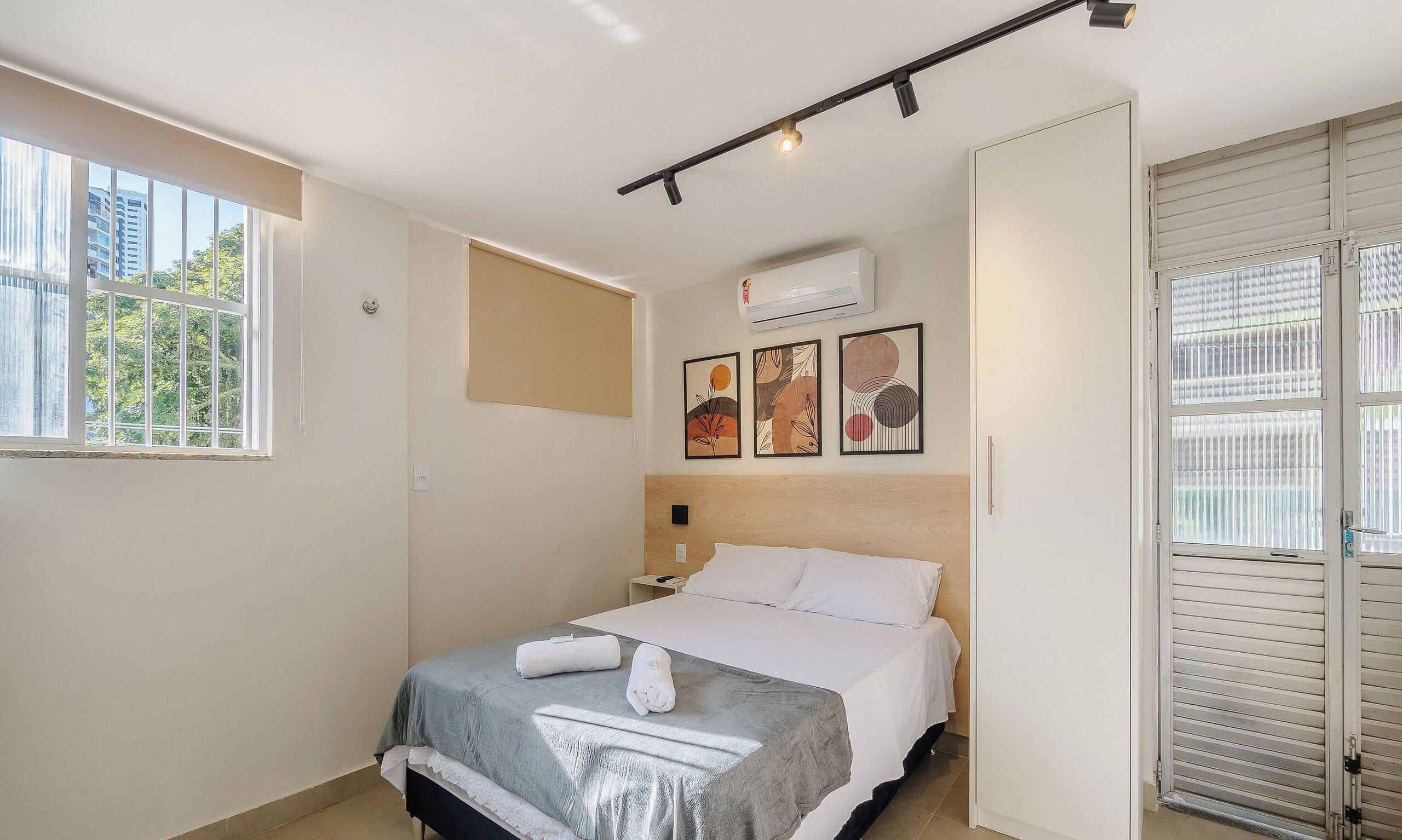 A hotel room with a bed, a window, an air conditioning unit, and a door with a view of the outside.