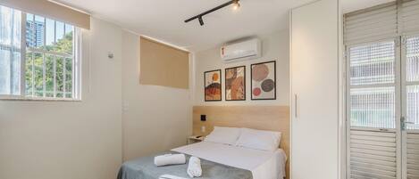 A hotel room with a bed, a window, an air conditioning unit, and a door with a view of the outside.