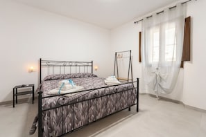 Een opgemaakt bed met een bedsprei met een patroon, een nachtkastje met een lamp en een raam met dunne gordijnen.