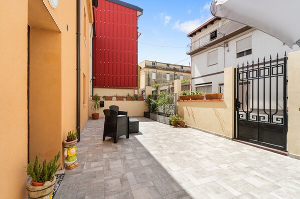 Une cour avec un sol carrelé, des meubles d’extérieur, des plantes en pot et une façade de bâtiment rouge.