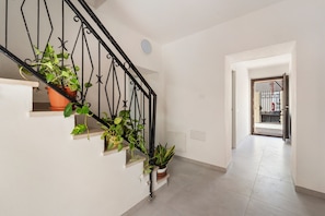 Un escalier orné de plusieurs plantes en pot, un mur blanc et un couloir menant à une porte surmontée d’un portail en métal.
