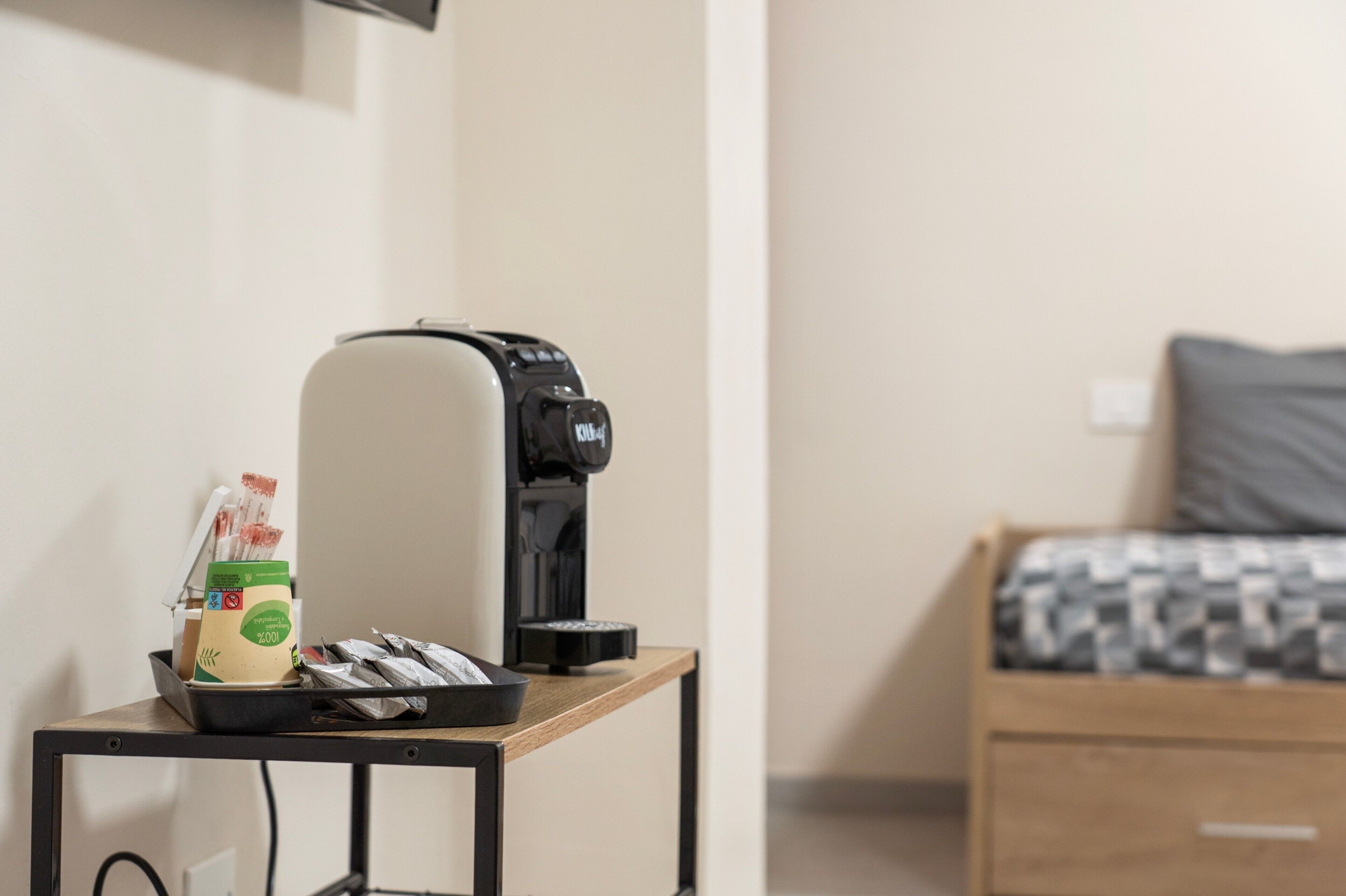 Ein kleiner Tisch mit Kaffeemaschine, einer Kaffeetasse und einigen Teebeuteln, ein Bett mit geblümtem Überwurf und eine schlichte Wand.