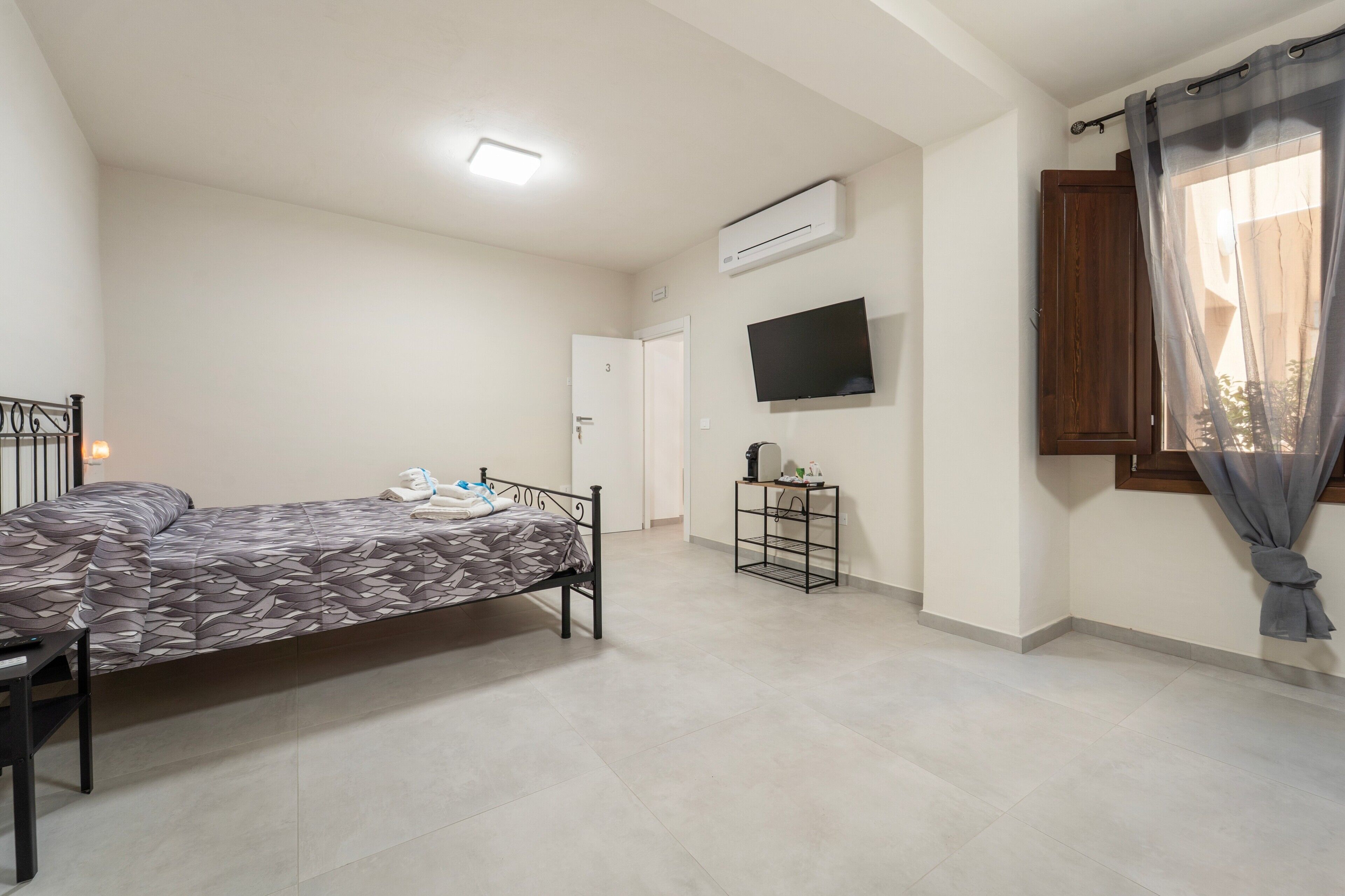 Un dormitorio con cama, televisor, aire acondicionado, puerta y ventana con cortinas.