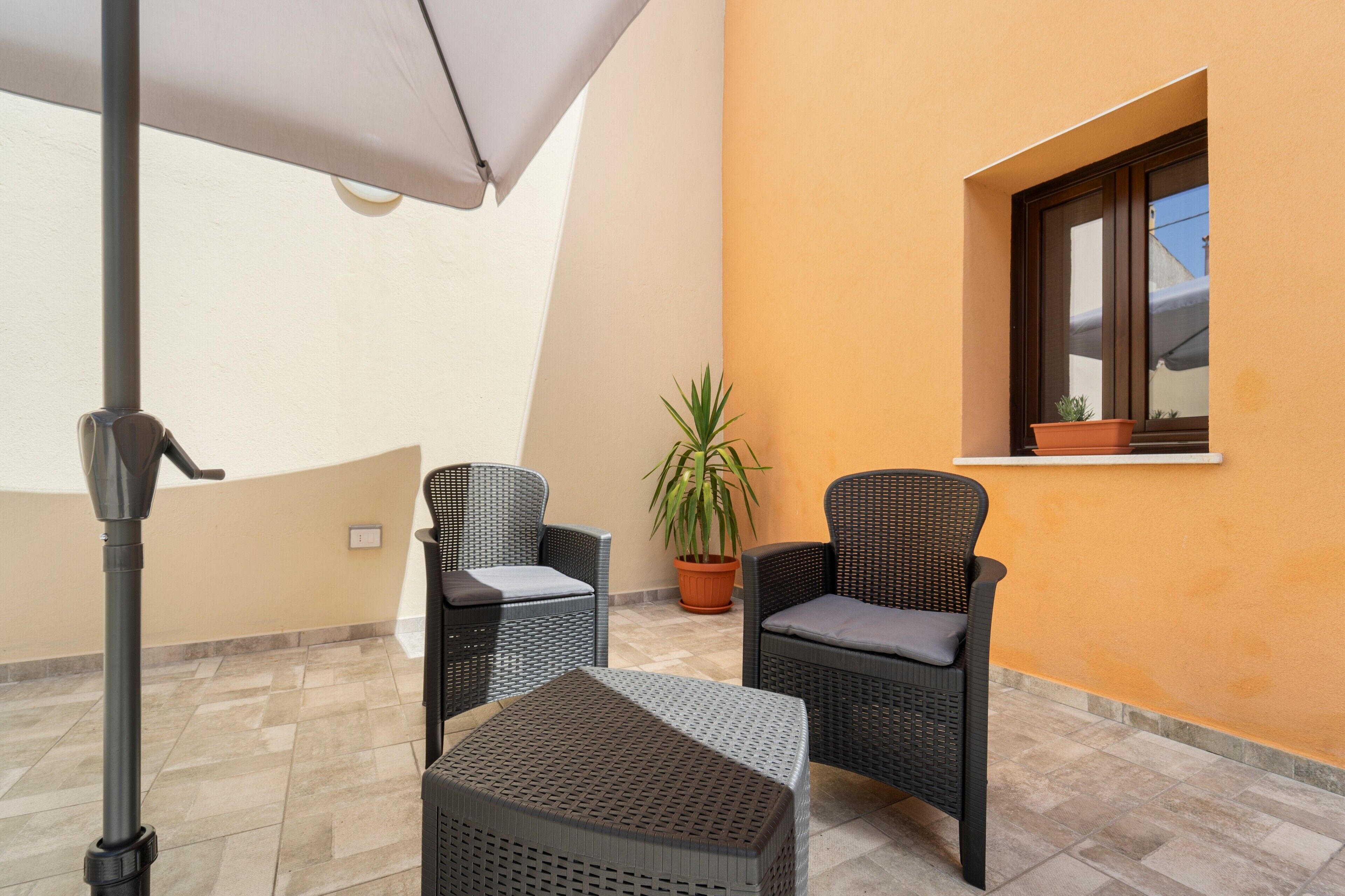Ein Balkon mit zwei Korbstühlen, einem kleinen Tisch, einem Blumentopf und einem Fenster mit einer Pflanze auf dem Sims.