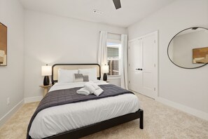 A neatly arranged bedroom with a bed, bedside lamps, a window with curtains, a door, and a round mirror on the wall. - Hawthorne Haven - 2.3 mi to DT-Direct Trail Access (Bentonville)
