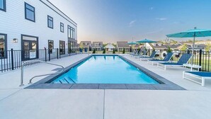 A swimming pool with lounge chairs and umbrellas, a white building with windows, and a clear sky.