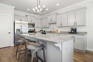 A modern kitchen with white cabinetry, a large island with bar stools, stainless steel appliances, and a chandelier.