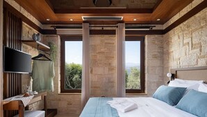 A bedroom with a large bed, a desk with a chair, a TV, and a window with a view of greenery.