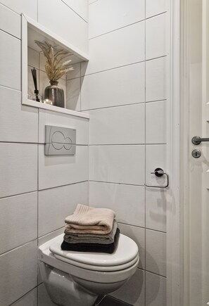 Ein Badezimmer mit weißen Fliesen, einer Toilette mit Stapel Handtüchern, einem Regal mit Vase und Kerzen sowie einer Tür mit Griff.