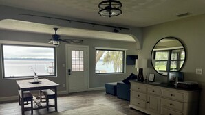 A spacious room with a wooden floor, a dining table with benches, a sofa, a dresser, and a large window offering a view of a body of water.