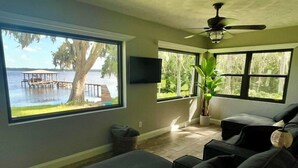 A living room with a large window overlooking a dock and water, a ceiling fan, and a flat-screen TV mounted on the wall.
