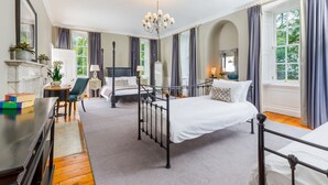 Un dormitorio con una cama con dosel, un escritorio con libros, una silla, una chimenea y ventanas grandes con cortinas.