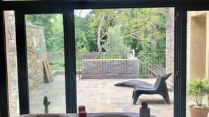 Een kamer met een glazen deur die leidt naar een terras buiten, met een stenen vloer, een zwarte lounge stoel en een cactus.