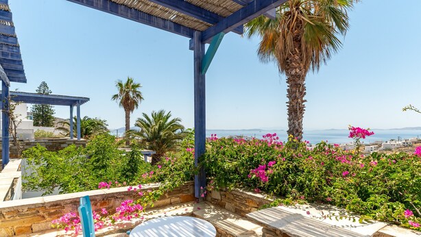 Una terraza con mesa y sillas, rodeada de muros de piedra y flores rosadas en flor, con vistas a la costa.