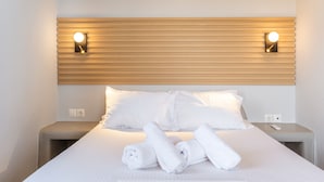 Habitación de hotel con cama, dos mesitas de noche, cabecera con luces de pared y toallas enrolladas sobre la cama.