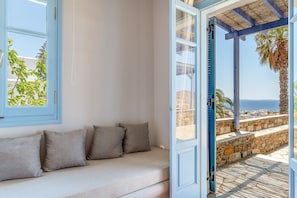 A room with a beige sofa, a window with blue shutters, and a view of the sea and palm trees. - Apartment 'Unit 1' with Sea View, Wi-Fi and Air Conditioning (Tinos)