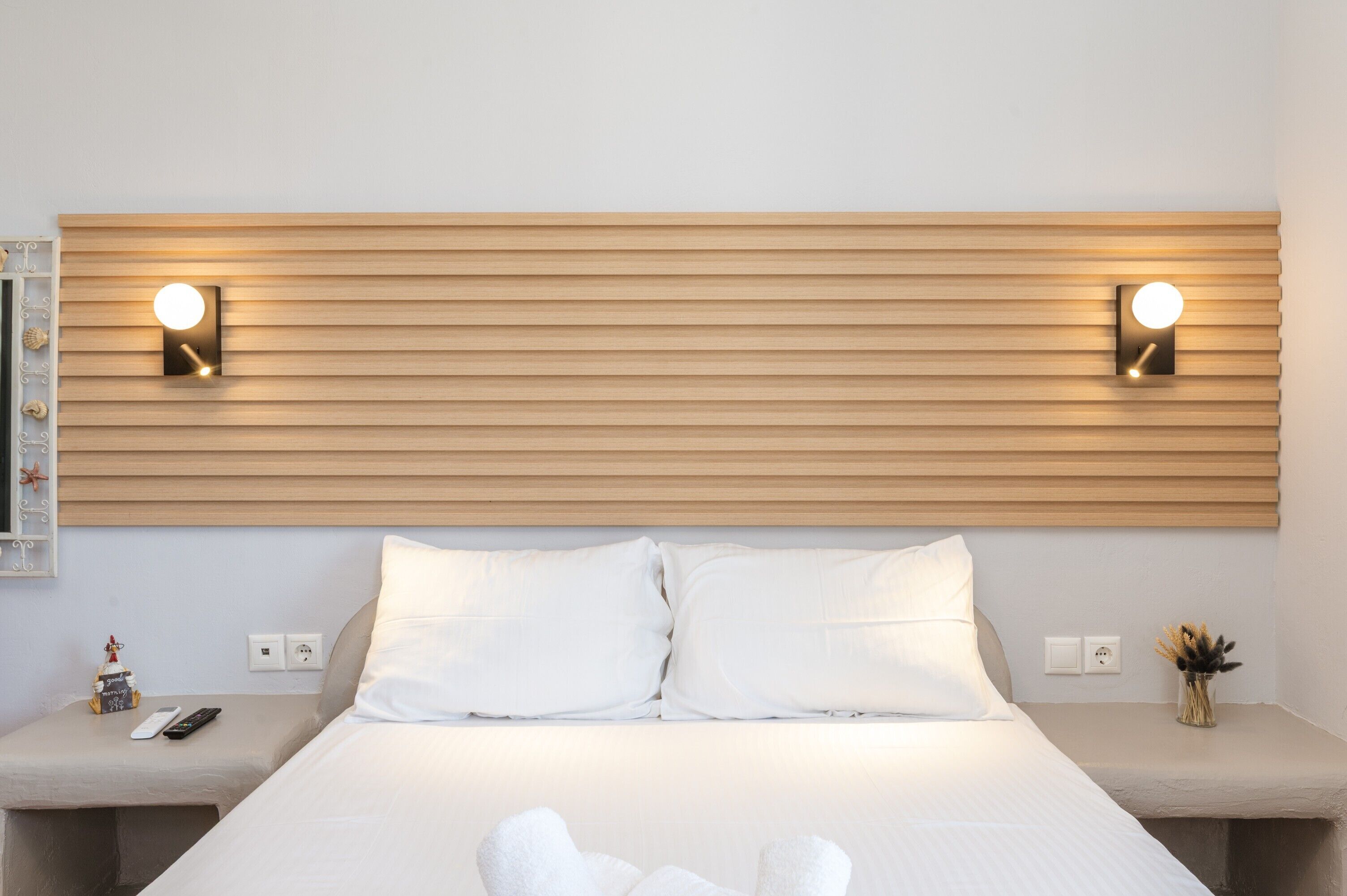 Une chambre d’hôtel avec une tête de lit en bois, deux lampes de chevet, un lit avec du linge de lit blanc, une table de chevet avec une télécommande et un vase contenant des fleurs séchées.