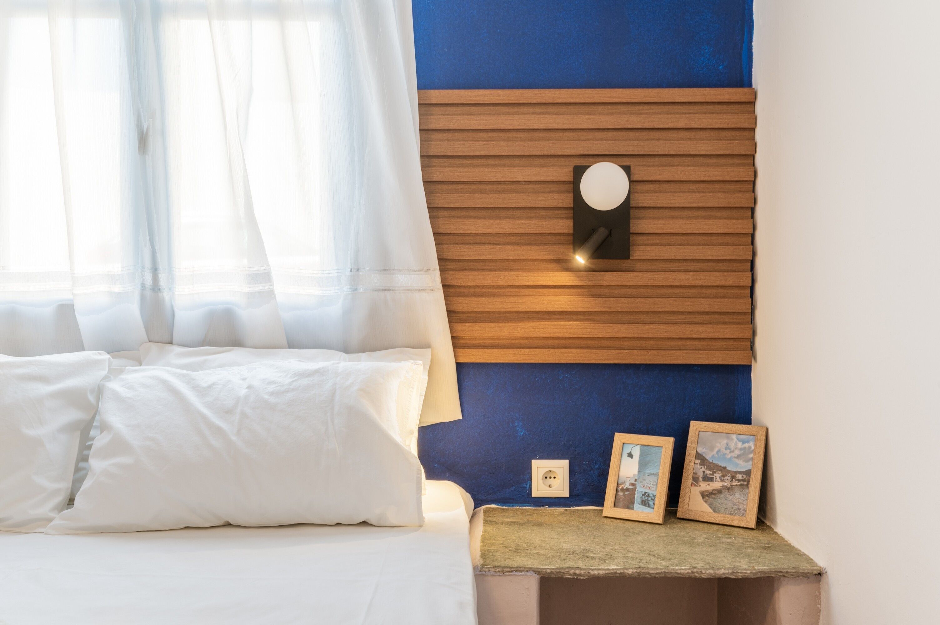 A hotel room with a bed, a nightstand with framed pictures, a wall-mounted light, and a window with sheer curtains.