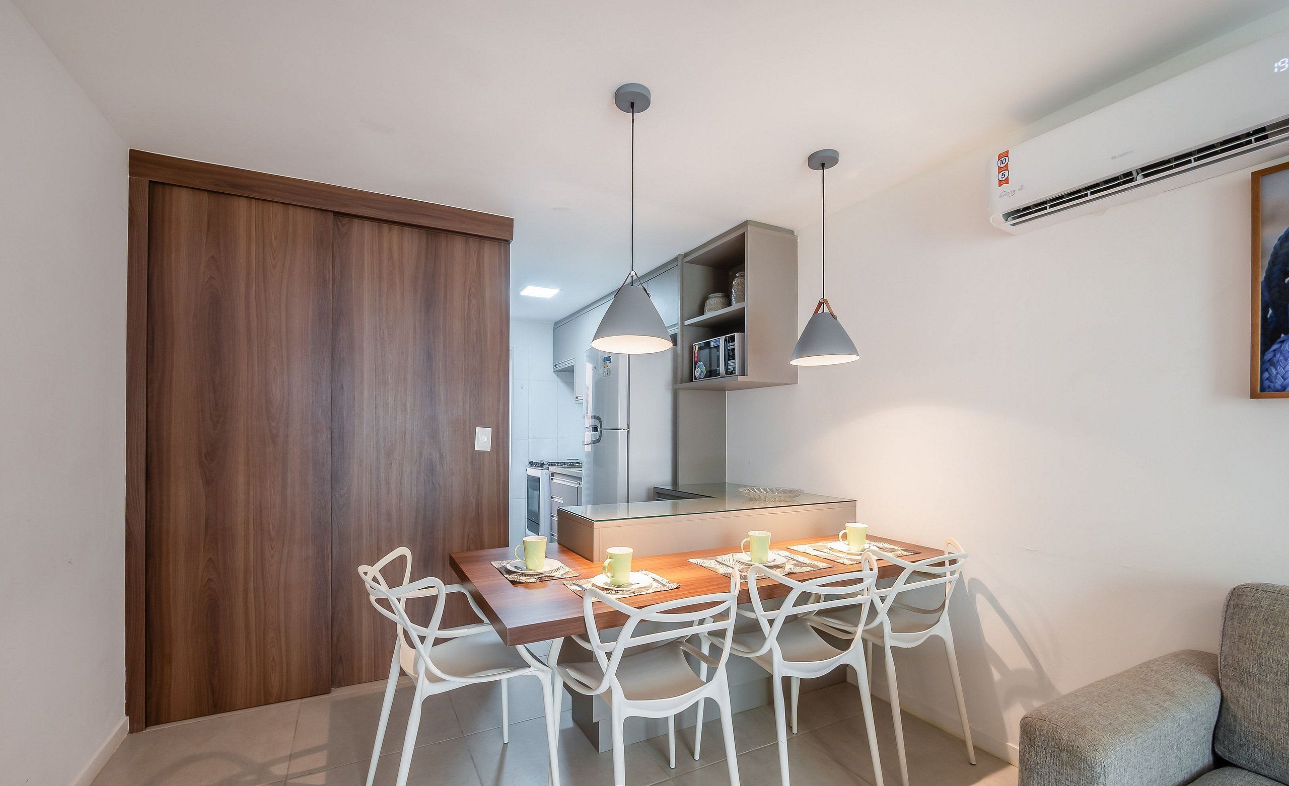 A modern kitchen with a dining area, white chairs, pendant lights, and a grey sofa.