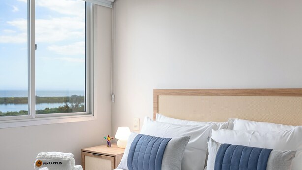 Un cuarto de hotel con cama, mesita de noche, lámpara, ventana con vista al mar y un equipo de aire acondicionado.