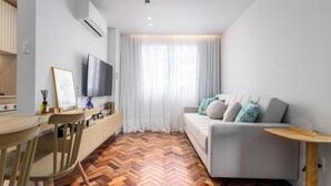 Una sala de estar moderna con piso de madera en patrón de espiga, un sofá gris con cojines estampados, una mesa de comedor de madera con sillas, un televisor de pantalla plana montado en la pared y un ventanal con cortinas translúcidas.