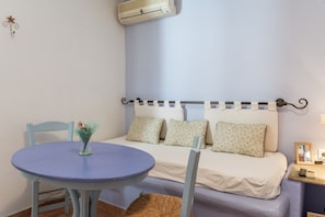 A room with a bed, a round table with a vase, a chair, a small bedside table with a picture and a phone, and an air conditioner on the wall. - Apartment '5' with Sea View, Wi-Fi and Air Conditioning (Tinos)