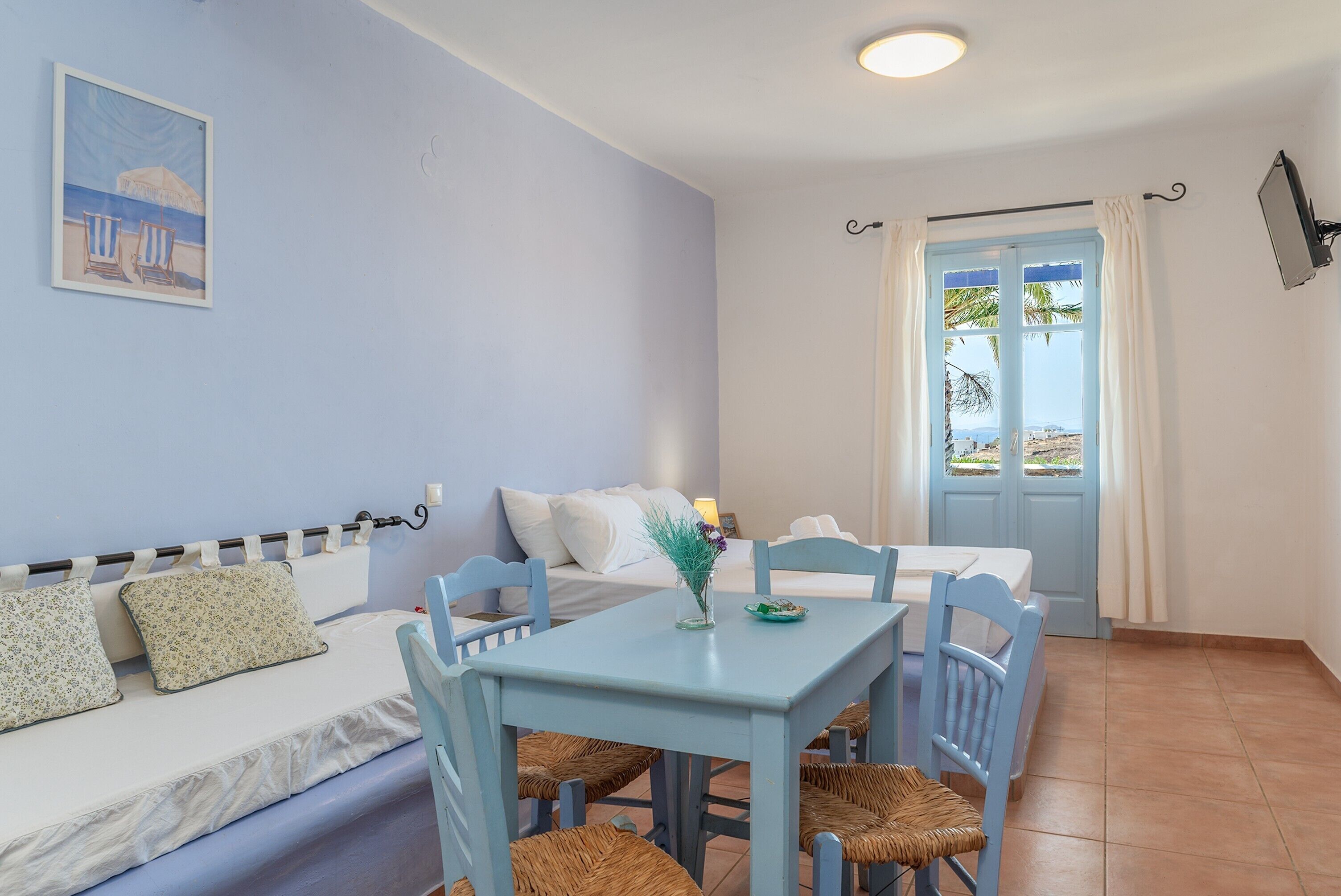 A room with a bed, a table with chairs, a painting on the wall, and a door leading outside.