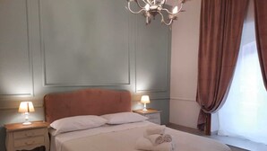 A neatly made bed with a headboard, two bedside tables with lamps, a chandelier, and a window with curtains. - Urbana collection guest house (Rome)