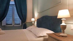 Una cama bien hecha con sábanas blancas, una mesita de noche de madera con una lámpara, y una ventana con cortinas azules.