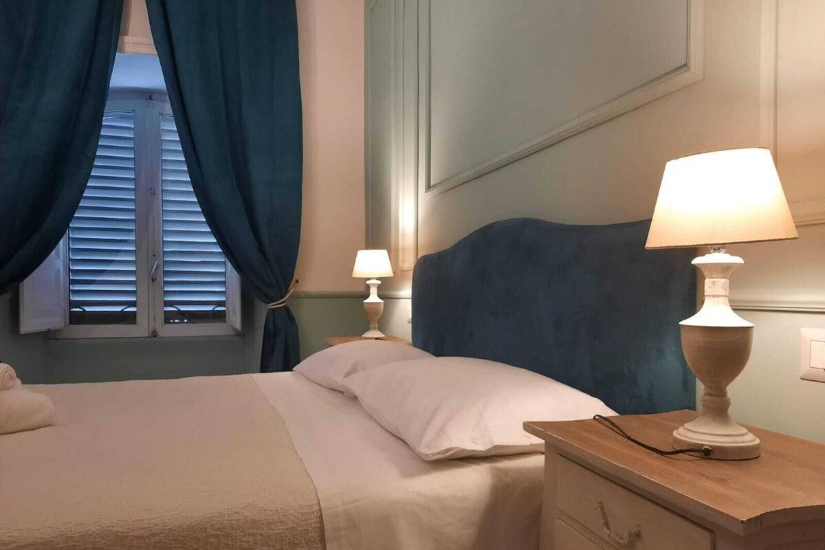 A neatly made bed with white linens, a wooden nightstand with a lamp, and a window with blue curtains.