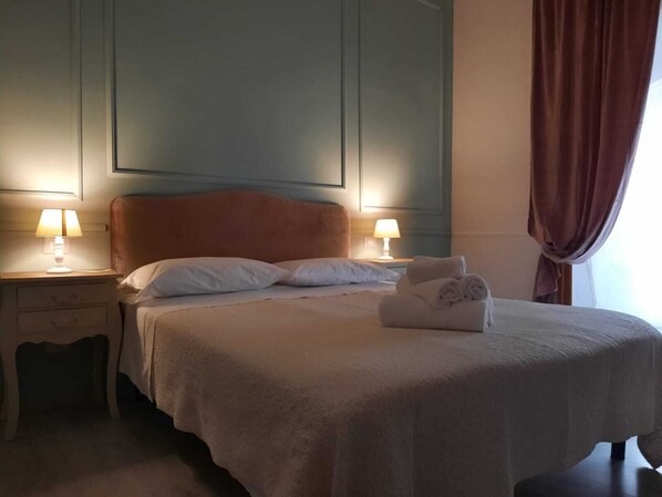 A neatly made bed with white linens and a folded towel on the side, two bedside lamps, a nightstand, and a window with curtains. - Urbana collection guest house (Rome)